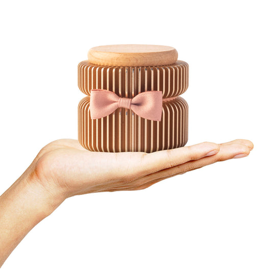 Keepsake urn held in palm for size reference – anodized aluminum with vertical stripes, beech wood lid, pink bow