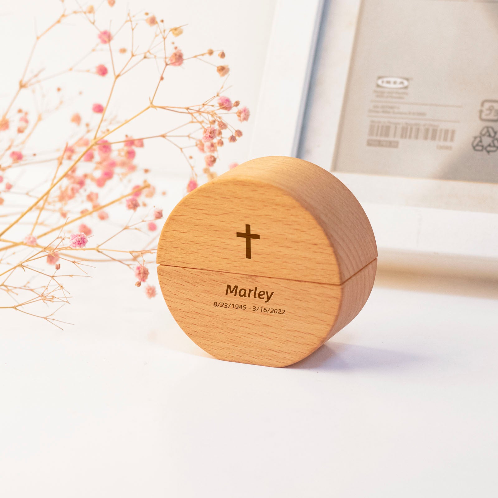 Round wooden keepsake urn with engraved cross, name "Marley", and dates "8/23/1945 – 3/16/2022" – displayed with dried flowers and frame, Christian memorial style